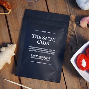 May include: A black pouch with white text that reads "The Satay Club" and "Life of Spice Gourmet Spice Blends". The text describes a versatile satay rub named after street food hawkers originally found close to Raffles Hotel in Singapore.