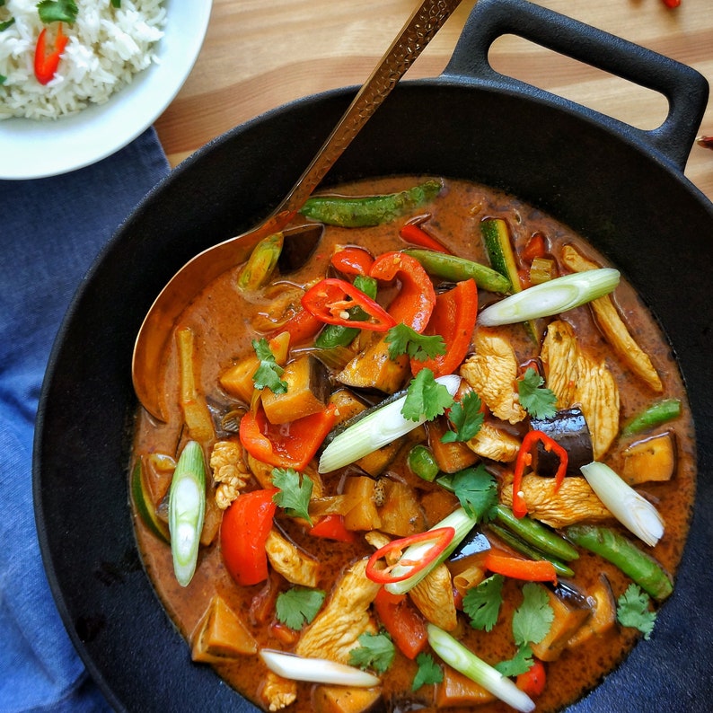 May include: A close-up of a black cast iron skillet filled with a red Thai curry. The curry contains chicken, eggplant, bell peppers, green beans, and scallions. The curry is garnished with cilantro and red chili peppers.