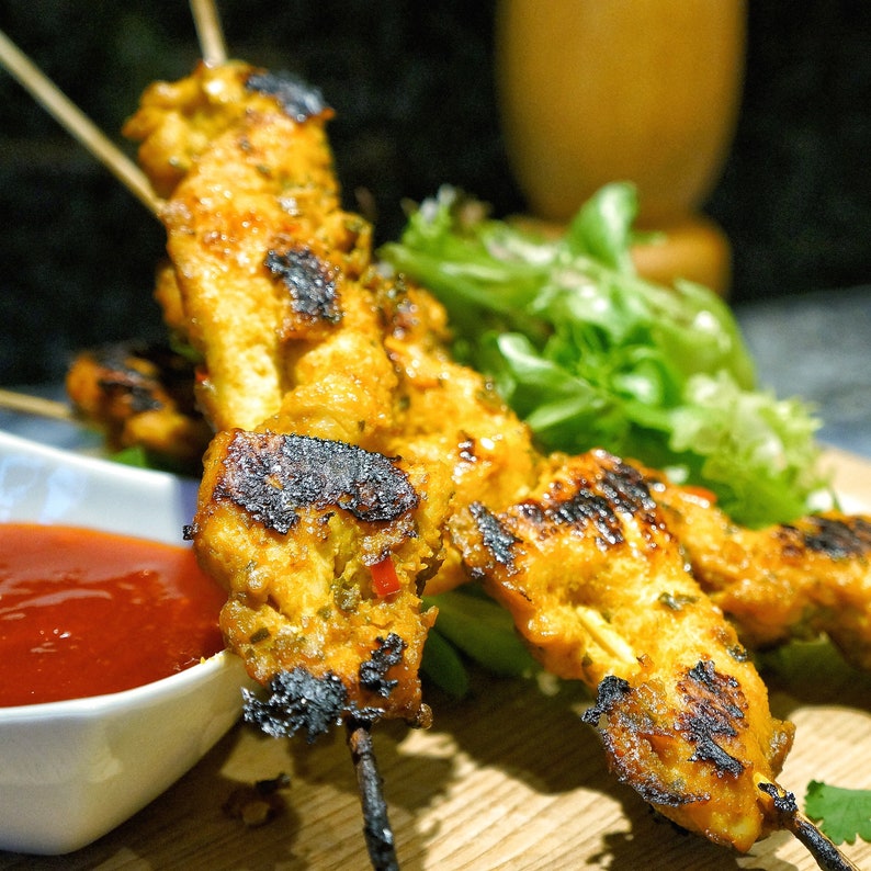 May include: Grilled chicken skewers with a yellow marinade and a side of green salad and a small bowl of red sauce.