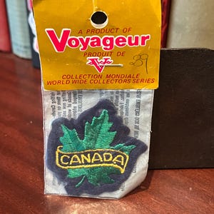 May include: A vintage Canada patch from the "Voyageur" World Wide Collectors Series. The patch features a green maple leaf and the word "CANADA" in yellow lettering on a dark blue background. The patch is still in its original packaging.