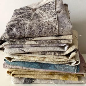May include: A stack of folded fabric squares featuring various textures and colours. The top pieces display a dark grey mottled pattern on a light background, followed by layers in solid muted tones of blue, yellow, and light grey with dark speckles.