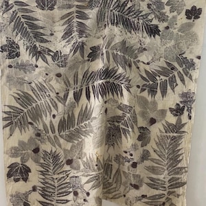 May include: A beige textile featuring a repeating pattern of dark grey leaf imprints. The leaves vary in size and shape, including ferns and broadleaf varieties. The fabric is suspended from wooden pegs.