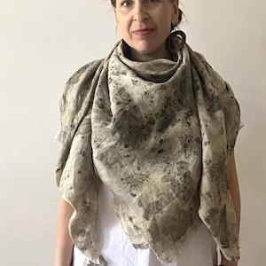 May include: A beige and olive green scarf with a leaf print. The scarf is draped over a white dress. The scarf has a natural, organic look, with a soft, flowing texture. The overall aesthetic is earthy and bohemian.