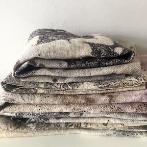 May include: A stack of folded rectangular fabric pieces featuring a textured, woven appearance. The fabric displays a mottled pattern in shades of dark brown, charcoal grey, and beige, with visible fibres and a coarse, natural-looking weave throughout.