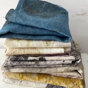 以下が含まれることがあります： さまざまな色のリネン生地が折りたたまれた山。最上層は青色で、クリーム色、タン色、マスタードイエローの層もあります。一部の生地には、濃い斑点模様があります。生地はきちんと積み重ねられており、その質感と色を示しています。