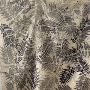 以下が含まれることがあります： クリーム色のテキスタイルで、濃い灰色のシダの葉の繰り返し模様があります。葉には目に見える葉脈があり、生地にプリントされています。木製の洗濯ばさみからテキスタイルが吊り下げられています。