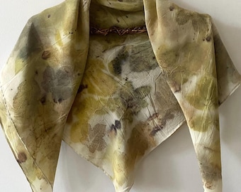 May include: A square scarf featuring a botanical print with various leaf shapes in shades of olive green, yellow, and brown. The fabric has a soft, slightly wrinkled texture and is draped over a twisted brown twig hanger against a plain wall.