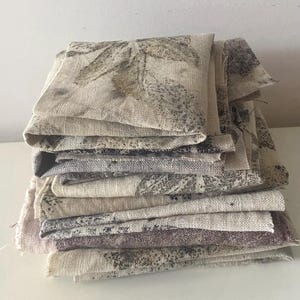 Op de afbeelding: Een stapel gevouwen vierkante stukken stof met een natuurlijke beige basiskleur en donkerbruine en grijze botanische bladprints. De stof heeft een zichtbare geweven textuur met gerafelde randen aan de zijkanten, wat een gelaagd uiterlijk geeft.