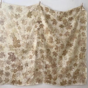 Pode incluir: Um tecido retangular grande com um padrão repetido de impressões de folhas de bordo em tons de castanho e bege. O tecido está pendurado num varal, mostrando o design natural das folhas. O efeito geral é rústico e orgânico.