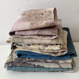 May include: A stack of folded fabric squares featuring a variety of textures and colours. The collection includes pieces with mottled brown and beige patterns, solid deep blue, and muted light blue tones, all displaying a visible woven textile weave.