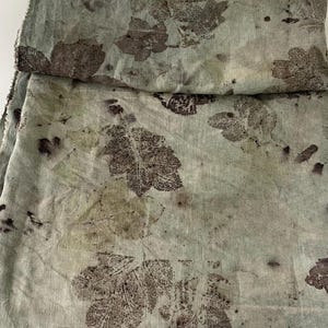 Puede incluir: Un trozo de tela doblado con un estampado de hojas verde claro y marrón. La tela tiene un borde crudo e inacabado. Las huellas de las hojas son en tonos marrones y están dispersas por la tela.