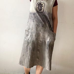 Könnte beinhalten: Eine Frau trägt ein graues Leinenkleid mit einem Batik-Muster und einem floralen Stickerei-Detail. Sie trägt schwarze Schnürschuhe und eine Halskette mit mehreren Perlensträngen.