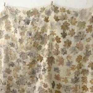 Pode incluir: Um tecido creme com um padrão de folhas de bordo em tons de castanho, cinza e bege. O tecido está pendurado com molas de roupa de madeira. As folhas estão espalhadas pela superfície, criando um design natural e orgânico.