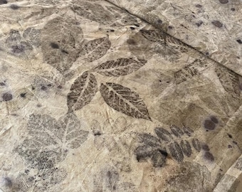 May include: A piece of light beige fabric featuring a botanical print with dark brown and grey leaf patterns. The textile displays a variety of detailed leaf shapes and dark circular spots scattered across the surface, with a folded edge visible.