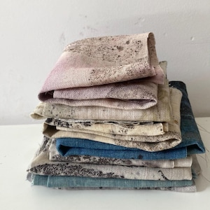 Pode incluir: Uma pilha de peças de tecido dobradas com um padrão texturizado e salpicado em várias cores, incluindo rosa suave, bege, castanho e tons de azul. O tecido tem um grão visível e parece ser linho ou uma fibra natural semelhante.