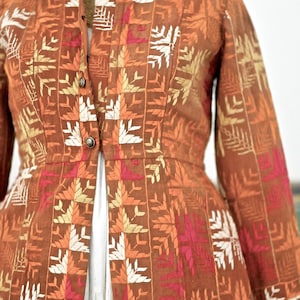 May include: A long, brown jacket with a mandarin collar and a button closure. The jacket is embroidered with a geometric pattern in white, orange, and pink.