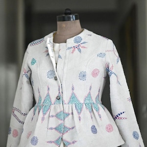 Könnte beinhalten: Eine weiße Jacke mit Schößchen, verziert mit einem sich wiederholenden Muster aus blauen, türkisfarbenen und rosa Stickereien. Die Jacke hat einen Stehkragen und wird vorne mit Knöpfen geschlossen. Das Kleidungsstück wird auf einer Büste präsentiert.