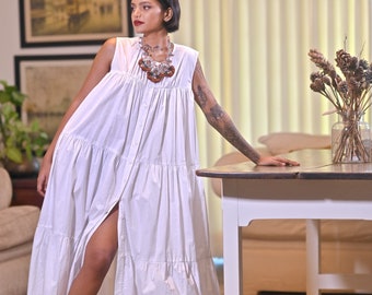 May include: A woman wearing a white sleeveless maxi dress with a tiered skirt and a brown and silver statement necklace. The dress has a button-up front and a slit in the skirt. She is standing in front of a white table with a vase of dried flowers.