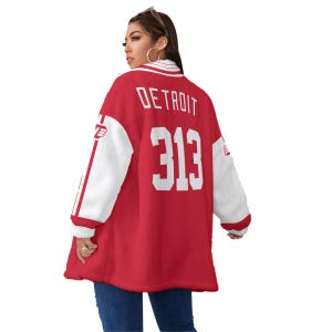 May include: A red and white letterman jacket with the word "DETROIT" and the number "313" on the back. The jacket has white sleeves and a white collar. The jacket is worn by a person.