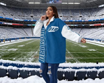 May include: A blue and white Detroit jacket with the word "DETROIT" printed vertically on the front. The jacket has a white fleece lining and a zipper closure. The jacket is worn in a snowy stadium.