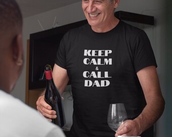 Puede incluir: Un hombre con una camiseta negra con el texto "Keep Calm & Call Dad" en letras blancas. Está sosteniendo una botella de vino y una copa de vino. Hay un pavo asado en primer plano.