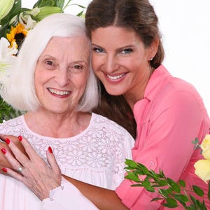 Puede incluir: Dos mujeres sonriendo y abrazándose, mirando a la cámara. Una mujer tiene el pelo blanco y lleva una blusa blanca con un diseño de encaje floral. La otra tiene el pelo castaño y lleva una camisa rosa abotonada. Hay flores en el fondo.