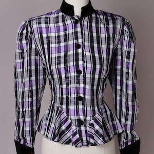 Könnte beinhalten: Eine langärmelige Jacke mit einem violetten, schwarzen und weißen Karomuster. Das Kleidungsstück verfügt über einen Stehkragen aus schwarzem Samt und passende Manschetten aus schwarzem Samt. Es hat eine Knopfleiste und einen Schößchensaum.