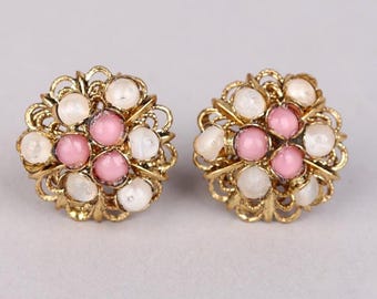 Puede incluir: Un par de pendientes de clip dorados. Cada pendiente presenta un diseño floral con cuentas rosas y blancas engastadas en un marco de filigrana. Los pendientes son redondos y tienen una estética vintage.