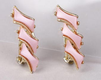 May include: A pair of gold-tone clip-on earrings featuring three stacked, wavy, light pink enamel segments. Each earring has a vertical, tiered design with smooth, curved edges and a polished metallic finish surrounding the pink inserts.