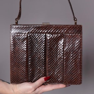 Peut inclure: Un sac à main marron vintage avec un motif en peau de serpent texturé. Le sac rectangulaire est doté d'une courte poignée et d'une fermeture à fermoir en métal. L'avant du sac présente des plis verticaux.