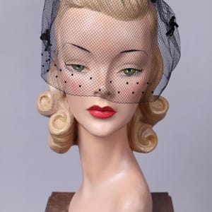 Puede incluir: Un velo negro de estilo vintage con diseño de red y pequeños adornos negros. El velo cubre una cabeza de maniquí con cabello rubio, ojos verdes y labios rojos. El maniquí se exhibe sobre un soporte de madera.