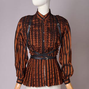 May include: A long-sleeved button-down shirt featuring vertical black and copper-brown stripes, a high collar, and vertical ruffles on the chest. The garment is accented with a black leather-style harness featuring thin straps and a waist belt.