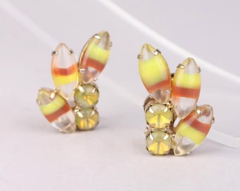 May include: A pair of gold-toned stud earrings. Each earring features a cluster of yellow, orange, and clear oval-shaped stones, arranged in a layered design. The earrings have a post-back closure and are set against a white background.