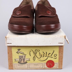 May include: A pair of brown suede shoes with a button closure. The shoes are sitting on a box with the word "Rinkle" written on it. The box also has a label with the text "BV" and "Artikel" on it.