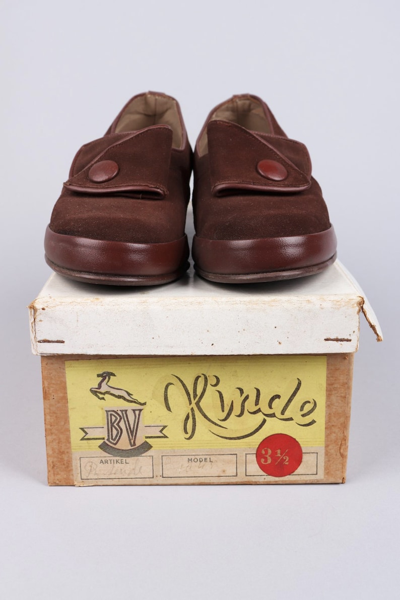 May include: A pair of brown suede shoes with a button closure. The shoes are sitting on a box with the word "Rinkle" written on it. The box also has a label with the text "BV" and "Artikel" on it.