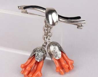 May include: A silver brooch featuring two coral-colored, flower-like pendants. The pendants are suspended from a silver chain and topped with silver caps. The brooch has a silver pin closure.