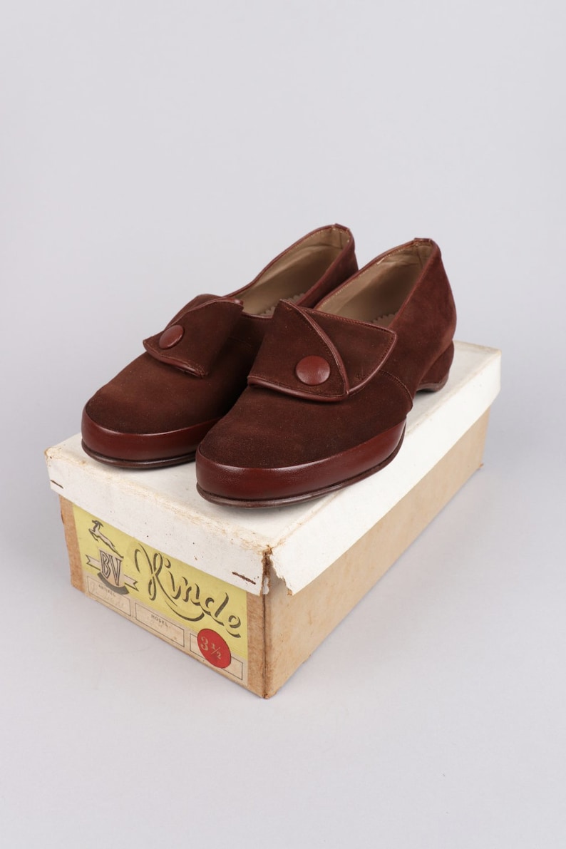 May include: A pair of brown suede shoes with a button detail on the top. The shoes are in a box with a yellow label that says "BV Kindle" and "3 1/2".