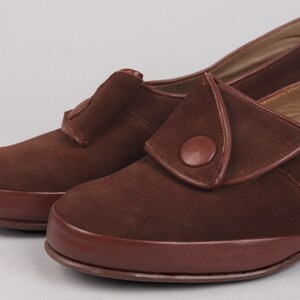 May include: A pair of brown suede shoes with a button detail on the strap. The shoes have a rounded toe and a low heel.