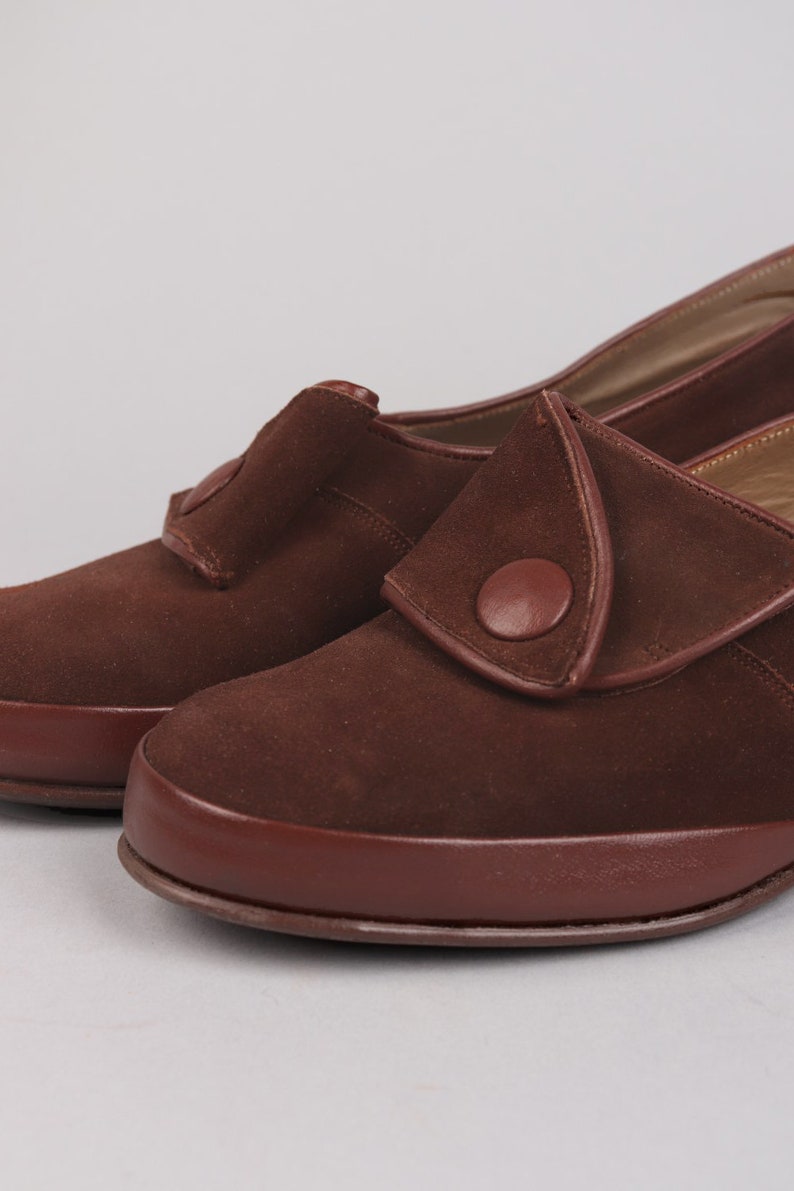 May include: A pair of brown suede shoes with a button detail on the strap. The shoes have a rounded toe and a low heel.