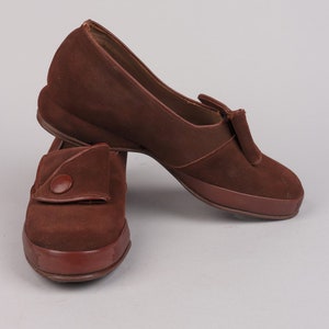 May include: A pair of brown suede shoes with a button detail on the front. The shoes have a low heel and a rounded toe.