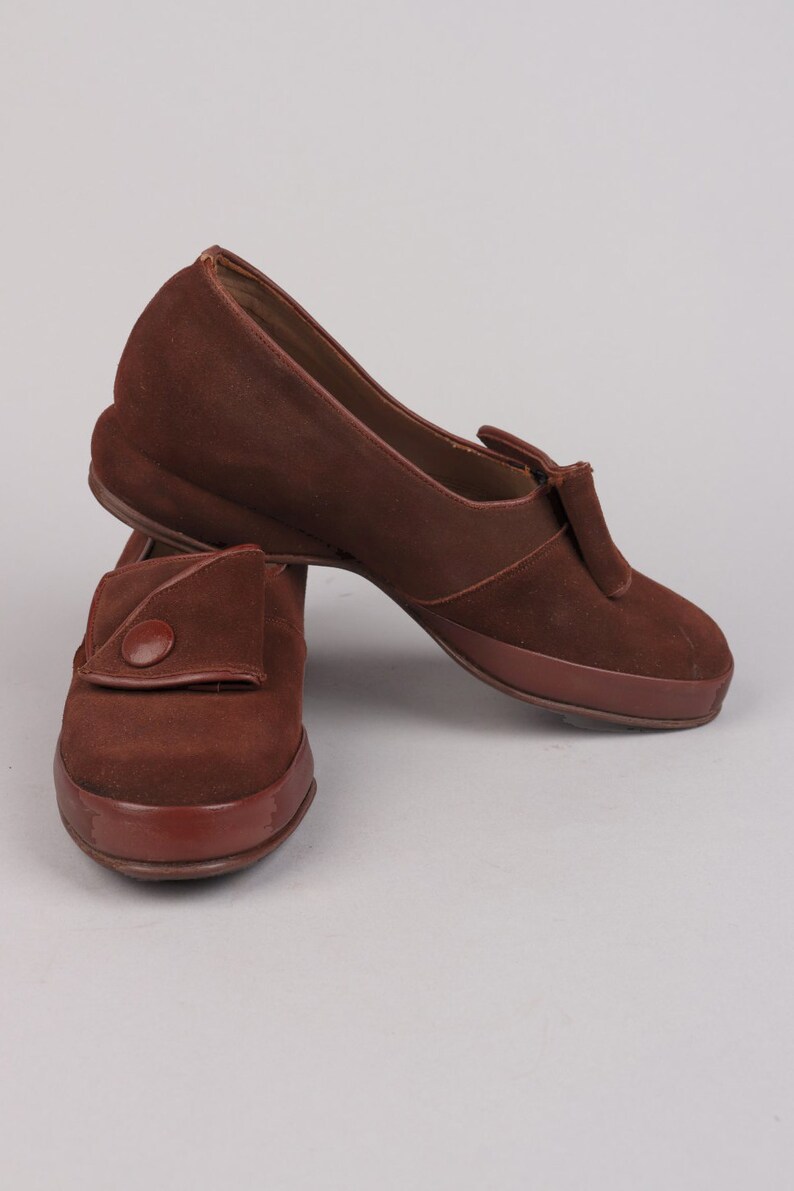 May include: A pair of brown suede shoes with a button detail on the front. The shoes have a low heel and a rounded toe.