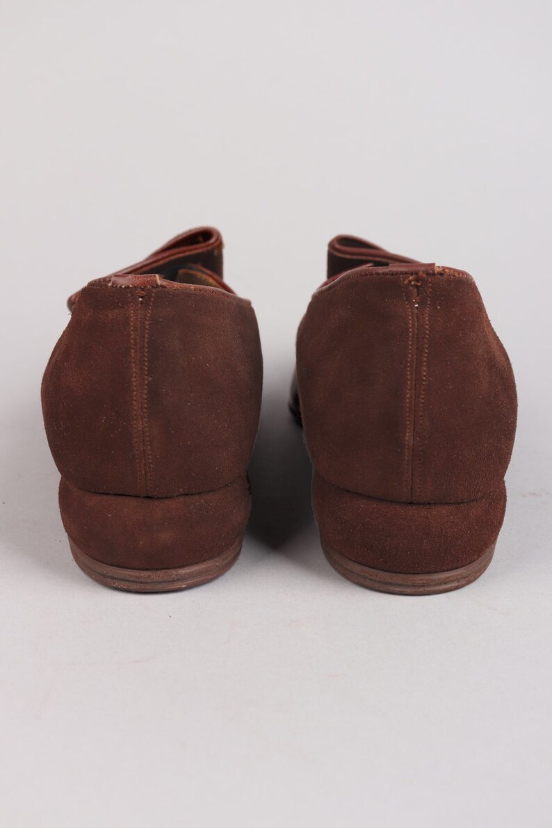 May include: A pair of brown suede shoes with a pointed toe and a low heel. The shoes have a strap across the top of the foot and a buckle closure.
