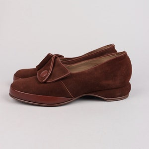 May include: Brown suede shoes with a button detail on the front. The shoes have a low heel and a rounded toe.