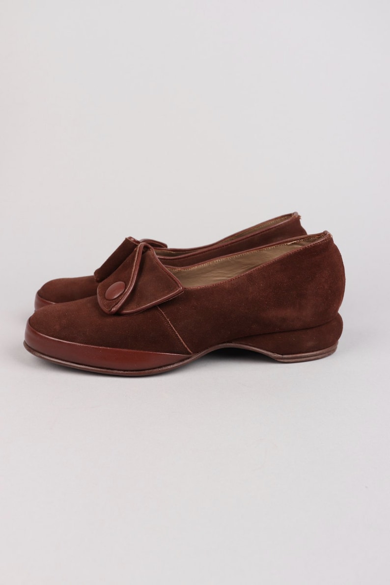 May include: Brown suede shoes with a button detail on the front. The shoes have a low heel and a rounded toe.