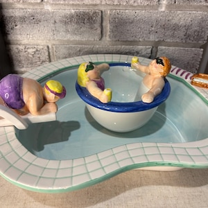 Könnte beinhalten: Eine Keramik-Servierschale in Form eines Schwimmbeckens mit drei Figuren. Eine Figur befindet sich auf dem Sprungbrett, zwei weitere im Becken. Das Becken hat einen blauen Innenraum mit einem weiß-grünen Kachelrand. Eine kleine Sandwich-Figur befindet sich an der Seite.