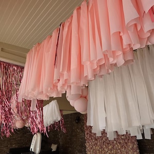 Könnte beinhalten: Festliche Außendekoration mit rosa und weißen Stoffbahnen, rosa Luftballons und metallischen rosa Fransen. Die Dekorationen hängen von einer weißen Decke, wodurch eine festliche Atmosphäre entsteht.