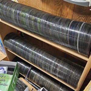 May include: A large collection of circular compact discs stacked horizontally on wooden shelving units. The discs are arranged in long, continuous rows, showing their reflective, iridescent edges. Some discs are stored in plastic cases below.