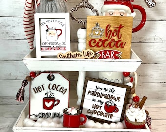May include: A white tiered tray decorated for a hot cocoa bar. The top tier features a gnome, a sign that says "The North Pole Hot Cocoa Co. Have a Cup of Cheer", and a wooden sign that says "Hot Cocoa Bar". The bottom tier features a sign that says "Hot Cocoa Served Here", a sign that says "You are the marshmallows in my hot chocolate", and a cupcake with whipped cream and a cinnamon stick.
