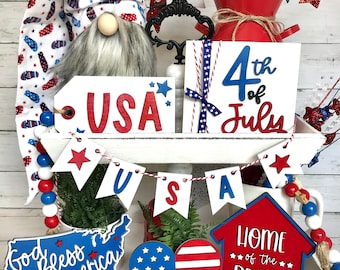 May include: A tiered white tray decorated with red, white, and blue patriotic themed decorations for the 4th of July. The decorations include a gnome wearing a red, white, and blue hat, a wooden sign that says "4th of July", a wooden sign that says "USA", a wooden sign that says "God Bless America", a wooden sign that says "Home of the Brave", a wooden heart with red, white, and blue stripes, and a red, white, and blue banner that says "USA".
