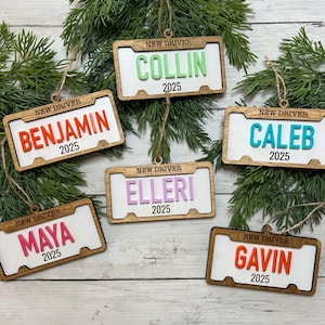 May include: Wooden license plate ornaments with names like Collin, Benjamin, and Caleb, in various colors. Each ornament has "NEW DRIVER" at the top and "2025" at the bottom. They are hanging on a green, leafy background.